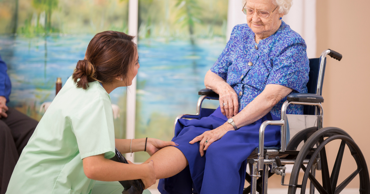 A female continuing care assistant helps an elderly female patient sitting in a wheelchair with her sore leg.