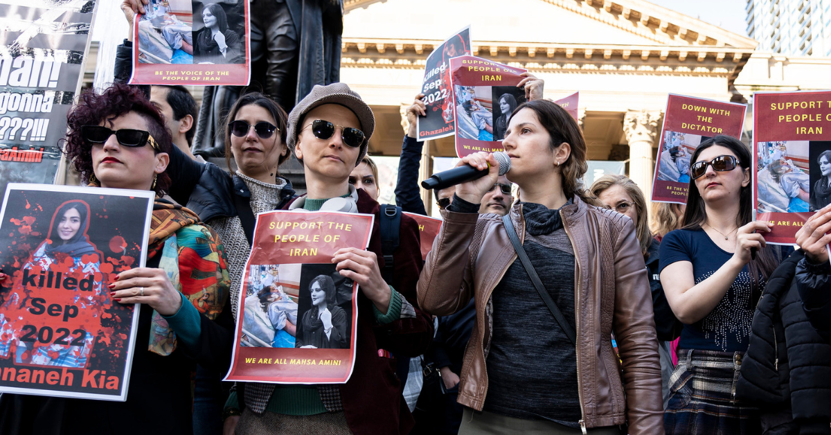 Photo of a group of women holding signs and speaking on a microphone at a solidarity protest with Iran