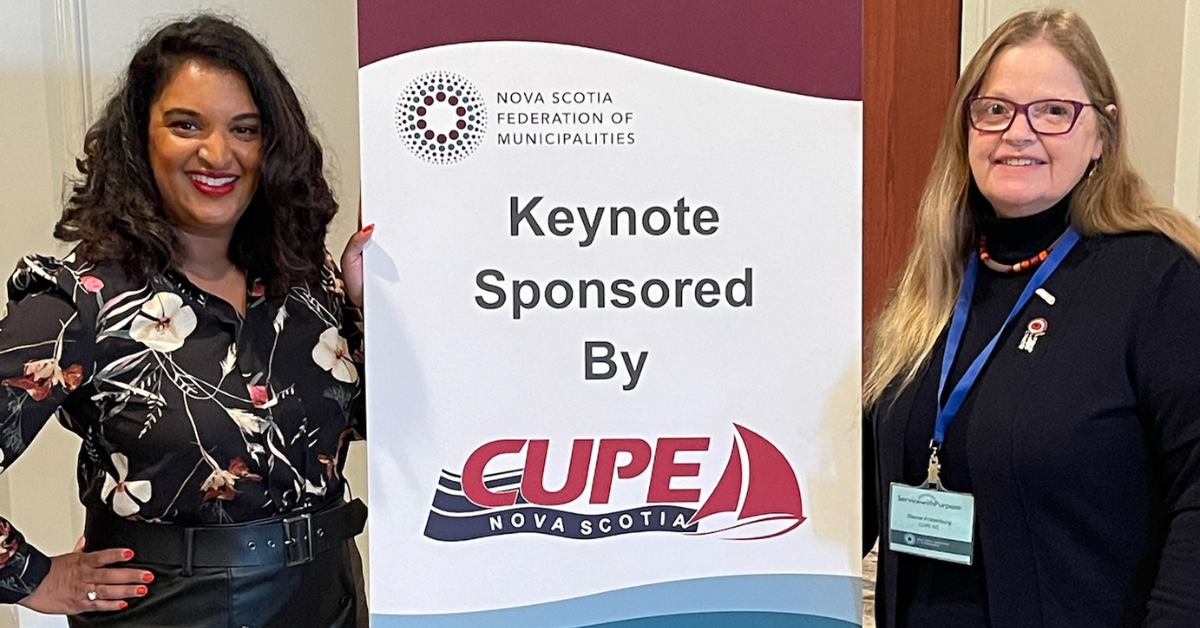 Tina Varughese and CUPE NS Vice-president Dianne Frittenburg, standing on either side of a sign that says, "Keynote speaker sonsored by CUPE Nova Scotia".